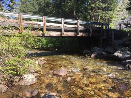 A wooden bridge spans a clear stream, surrounded by lush greenery and rocky terrain under a blue sky.
