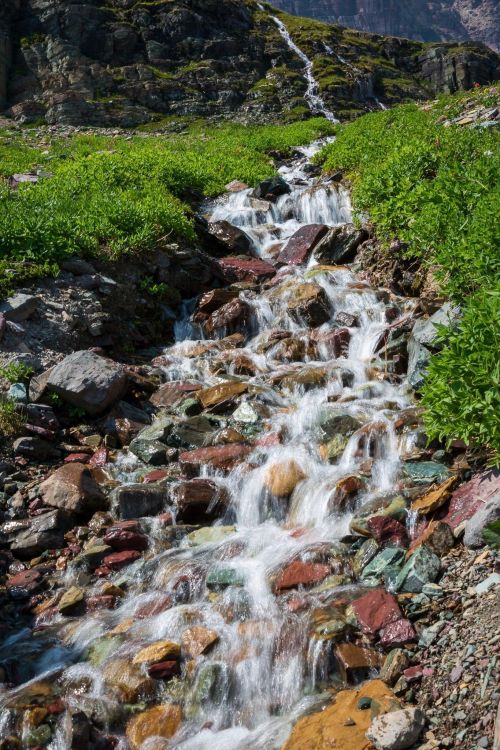 A vibrant stream flows over colorful rocks, surrounded by lush green vegetation and rocky terrain.