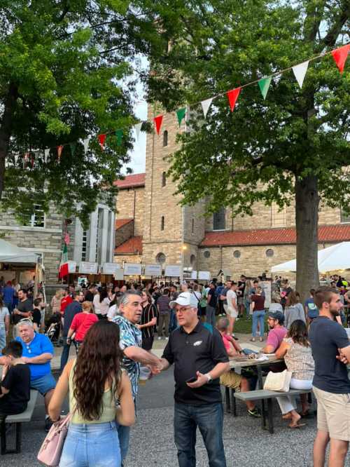 A lively outdoor festival scene with people mingling, food stalls, and festive decorations under trees.