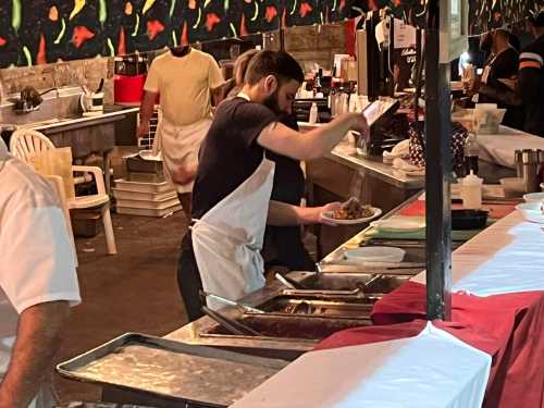 A chef in an apron serves food on a plate in a busy kitchen with other staff and cooking equipment visible.