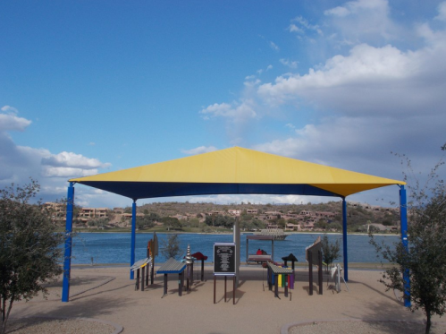A colorful shaded area with benches by a lake, surrounded by shrubs and under a partly cloudy sky.