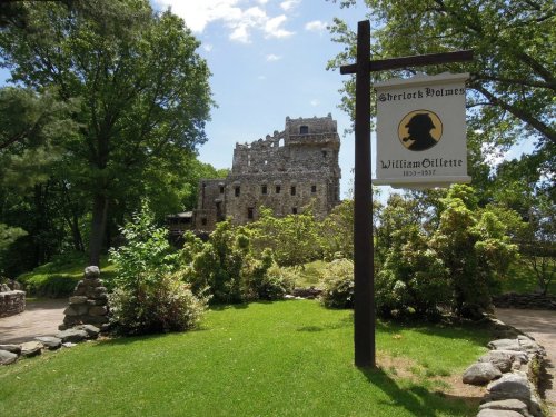 A stone castle ruins surrounded by greenery, with a sign for "Sherlock Holmes" and "William Gillette" in the foreground.
