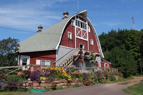 A red barn with a unique roof design, surrounded by colorful flowers and greenery under a clear blue sky.