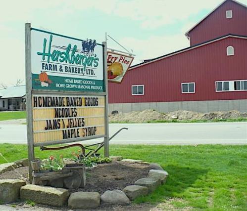 Sign for Hershberger's Farm & Bakery listing homemade baked goods, noodles, jams, jellies, and maple syrup. Red barn in background.