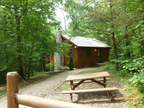 A wooden cabin surrounded by trees, with a picnic table in front and a gravel path leading to the entrance.