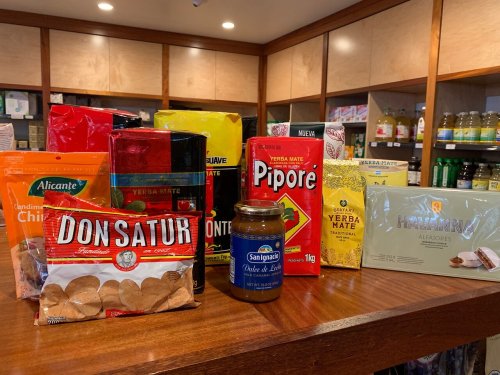 A variety of packaged foods and drinks, including yerba mate, cookies, and a jar of dulce de leche, on a wooden shelf.