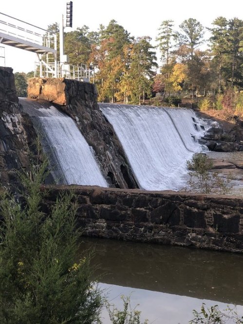 A waterfall cascading over a rocky dam, surrounded by trees and a calm body of water below.
