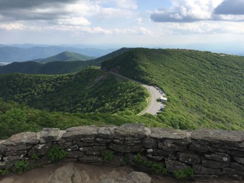 A scenic view of rolling green hills and a winding road, with a stone wall in the foreground under a cloudy sky.