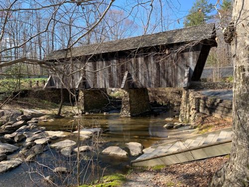 A wooden covered bridge spans a small stream, surrounded by trees and rocky terrain on a clear day.