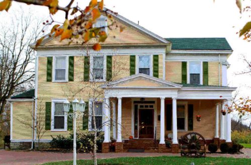 A large, yellow Victorian-style house with green shutters and a front porch, surrounded by trees and autumn leaves.