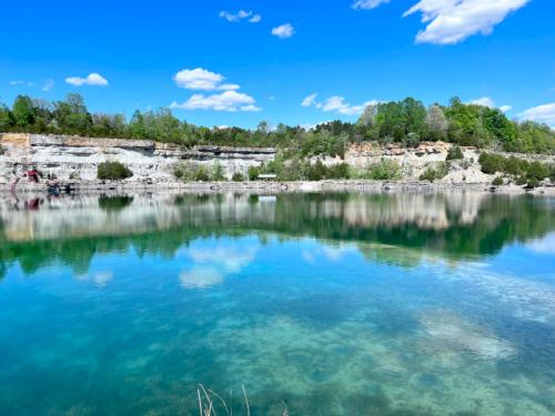 A serene quarry lake surrounded by lush greenery and rocky cliffs under a bright blue sky with fluffy clouds.