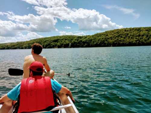 Two people in a canoe on a lake, surrounded by green hills and a blue sky with fluffy clouds.