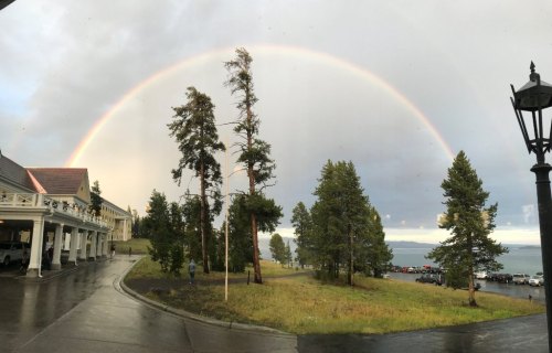 A vibrant rainbow arcs over a scenic landscape with trees, a building, and a lake under a cloudy sky.