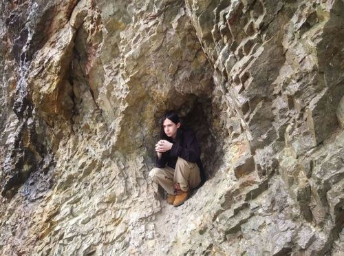 A person sits in a rocky cave, hugging their knees, surrounded by textured stone walls.
