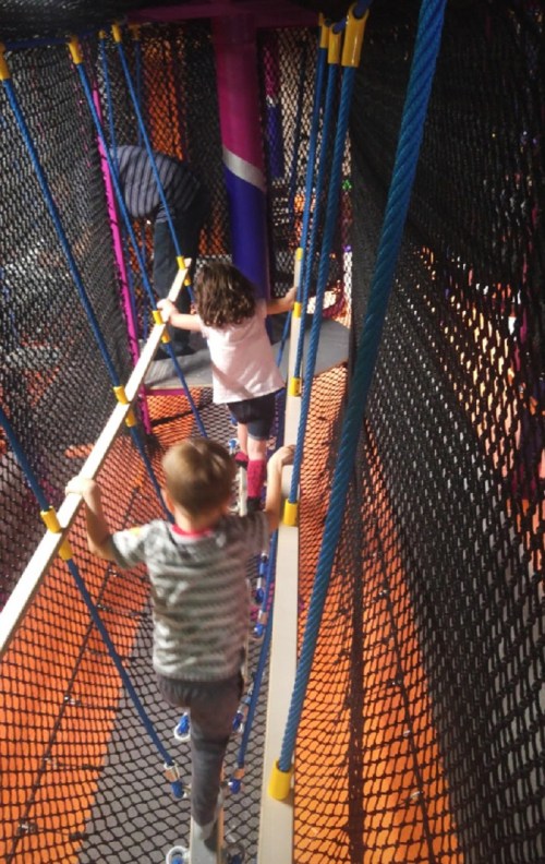 Two children navigate a rope bridge in a colorful indoor play area, surrounded by nets and vibrant structures.