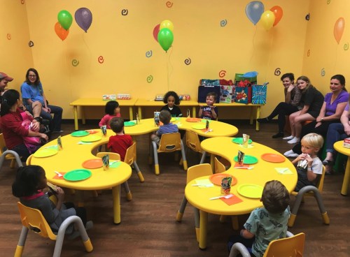 A colorful party room with children seated at yellow tables, balloons overhead, and adults watching from the sides.