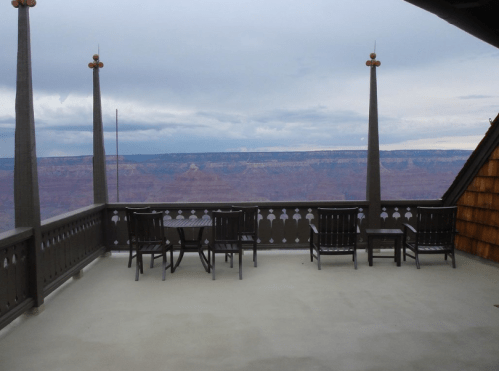 A balcony with wooden furniture overlooks a vast canyon under a cloudy sky.