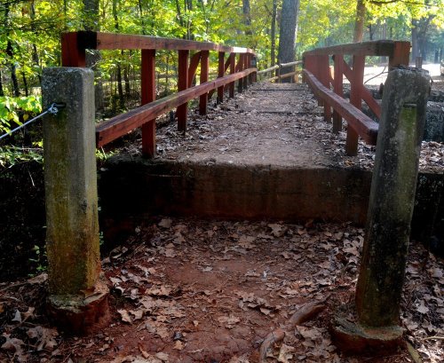 A wooden bridge with railings spans a path covered in fallen leaves, surrounded by trees in a forested area.