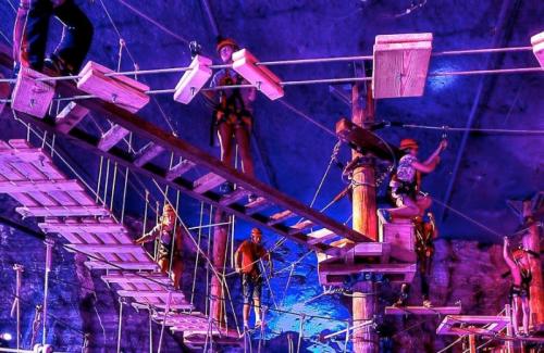 A group of people navigating a high ropes course with wooden platforms and safety gear, illuminated by colorful lights.
