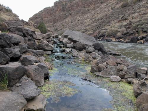 A rocky riverbank with a small stream flowing over stones, surrounded by dry hills and sparse vegetation.