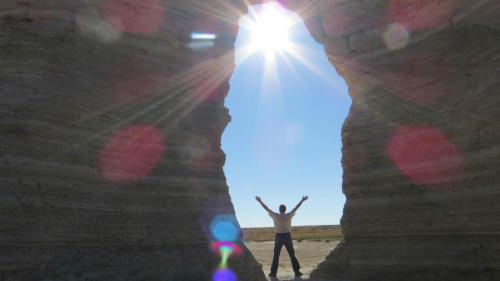 A person stands with arms raised in a rock formation, sunlight shining through the opening above.