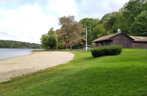 A serene lakeside scene featuring a sandy beach, green grass, and a rustic cabin surrounded by trees.
