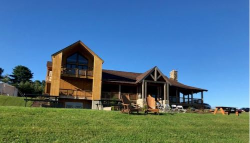 A large wooden house with a sloped roof, surrounded by green grass and clear blue skies.