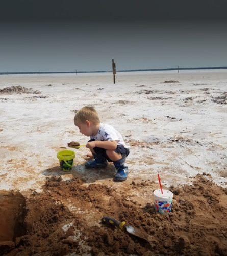 A young child squats on a sandy surface, playing with a green bucket and a cup, with a shovel nearby.