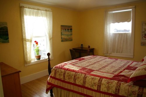 Cozy bedroom with yellow walls, a bed with a red and white quilt, a desk, and a window with a potted plant.