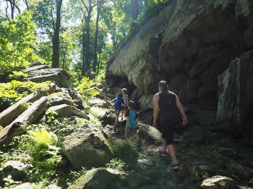 A woman and two children hike up a rocky trail surrounded by lush greenery and trees.