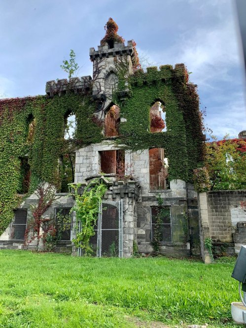 Ruins of a stone building covered in ivy, with broken windows and a tower, set against a cloudy sky.