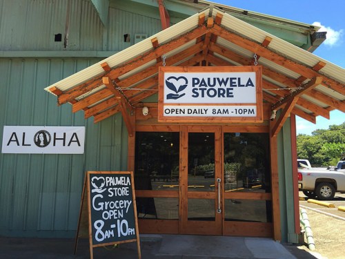 Entrance of Pauwela Store with a sign stating open hours, surrounded by a green building and a "Aloha" sign.