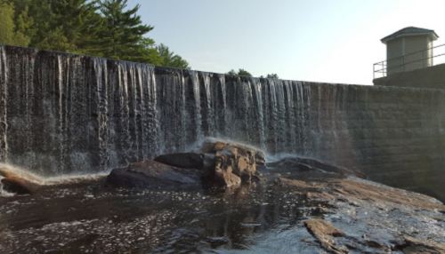 A serene waterfall cascading over a stone dam, surrounded by lush greenery and a clear blue sky.