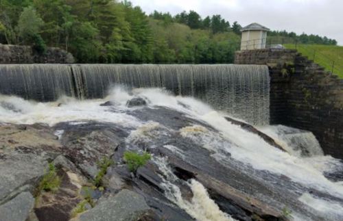A waterfall cascading over a stone dam, surrounded by lush greenery and rocky terrain.