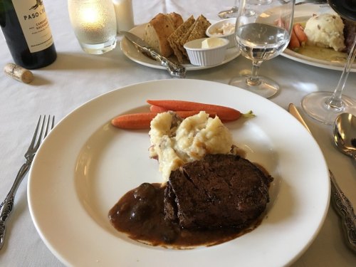 A plate with a beef steak in sauce, mashed potatoes, and carrots, accompanied by wine and bread on the table.
