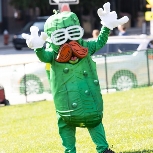 A person in a green pickle costume with a mustache and sunglasses, waving cheerfully in a park.