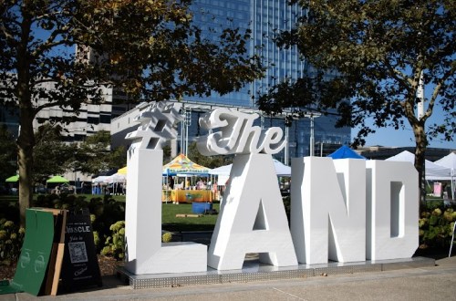 Large white letters spelling "The LAND" in a park with tents and trees in the background.