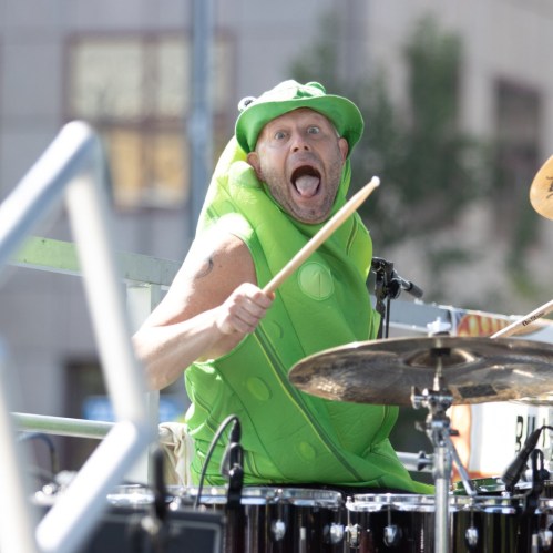 A drummer in a green costume and hat enthusiastically plays the drums, making a playful expression.