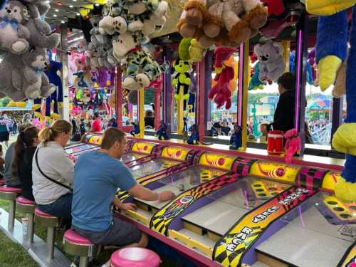 A carnival game booth with players trying to win stuffed animals, colorful prizes hanging above, and a festive atmosphere.