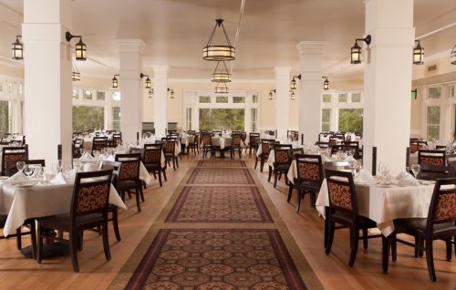 Elegant restaurant interior with tables set for dining, featuring wooden chairs and large windows for natural light.