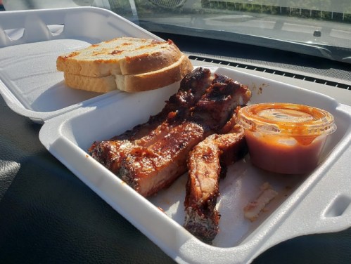 A takeout container with grilled ribs, a side of sauce, and two slices of bread on a car dashboard.