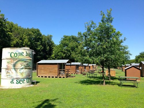 A grassy area with wooden cabins and a large water tank that says "Come back & see us" under a clear blue sky.