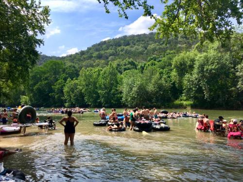 A crowded river scene with people floating on inner tubes and relaxing on the shore, surrounded by lush greenery.