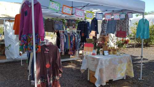 A colorful market stall with clothing hanging and a table displaying handmade items under a white canopy.