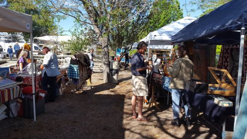 A vibrant outdoor market scene with vendors and shoppers browsing colorful stalls under sunny skies.