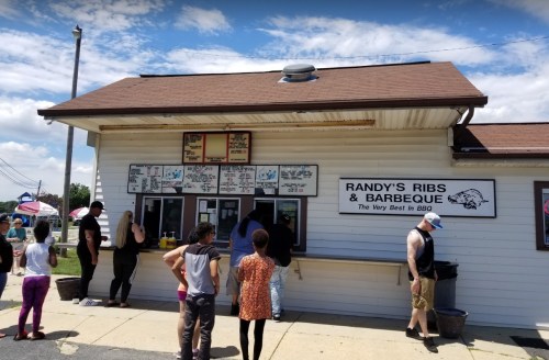 A busy BBQ stand with a menu sign, people in line, and a clear blue sky in the background.