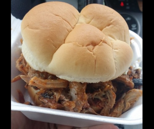 A close-up of a pulled pork sandwich with two soft buns, held in a takeout container.