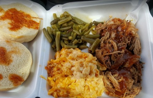 A takeout container with pulled pork, macaroni and cheese, green beans, and two bread rolls with sauce.
