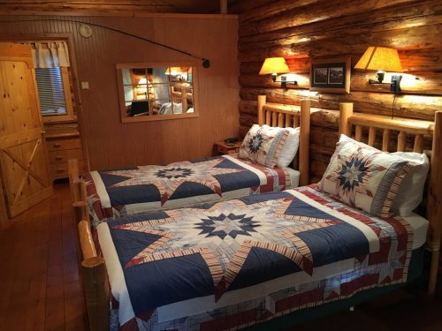 Cozy log cabin interior with two beds featuring star-patterned quilts, warm lighting, and rustic wooden decor.