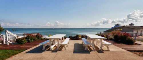 A serene waterfront view featuring white picnic tables overlooking a calm lake and distant boats under a blue sky.
